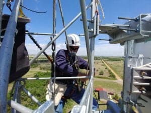 Technician in a fall-arrest harness and hard hat working near the top of a communications tower